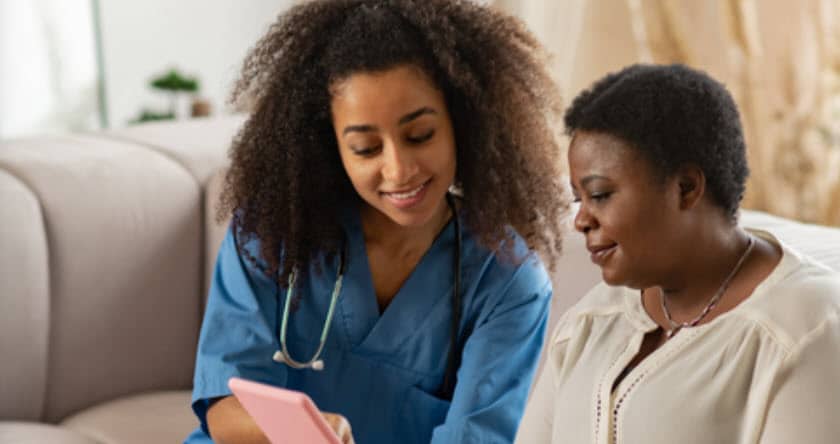Pleasant nurse. Pleasant curly nurse wearing blue uniform showing online news to elderly African-American lady